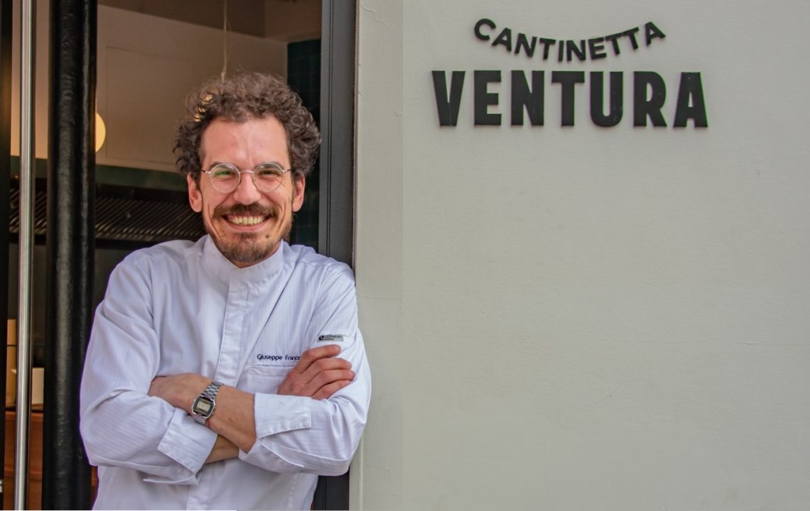 serge ventura, moustachu châtain bouclé, adossé à l'enseigne de son restaurant Cantinetta Ventura