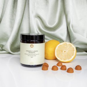 L'image montre un pot de gummies à la vitamine C au citron, posé à côté de citrons entiers et coupés, avec quelques gummies dispersés sur une surface blanche.