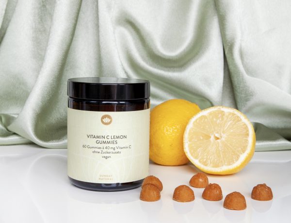 L'image montre un pot de gummies à la vitamine C au citron, posé à côté de citrons entiers et coupés, avec quelques gummies dispersés sur une surface blanche.