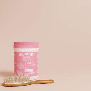 L'image montre un pot de Vital Proteins Beauty Collagen à côté d'une brosse à cheveux en bois sur une surface rose pâle.