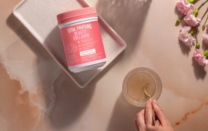 L'image montre un pot de Vital Proteins Beauty Collagen posé sur un plateau, à côté d'une main tenant une tasse de boisson, avec des fleurs roses en arrière-plan.