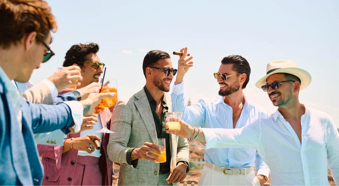 Groupe d’hommes élégants en costumes d’été Hockerty trinquant sur un rooftop à Florence pendant le Pitti Uomo 108.